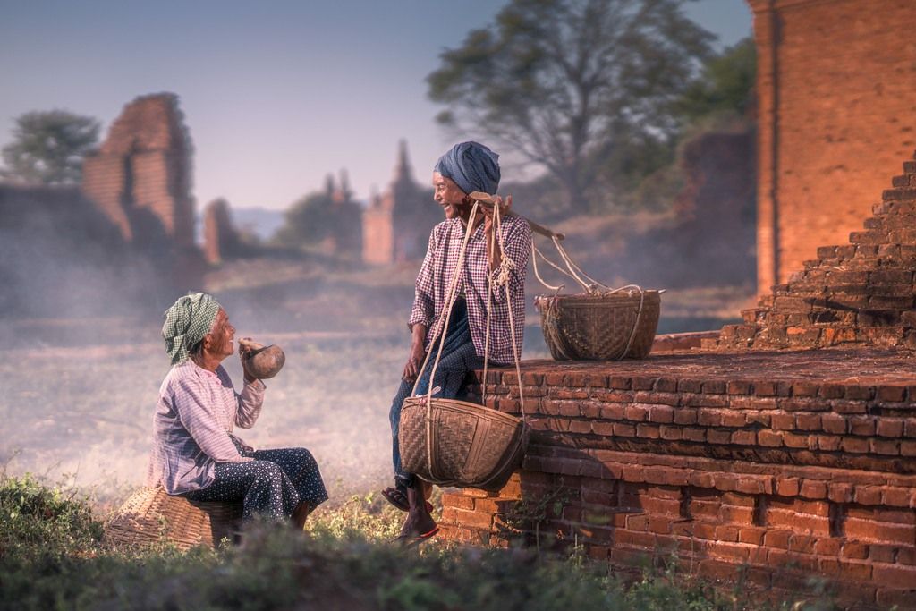 Two Old Women Sitting On The Benches Drinking Something Showing A Scene Of An Village Hometown